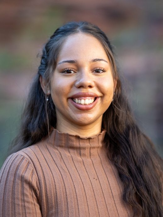 A young woman with long brown hair, wearing a ribbed brown top, smiles at the camera. The background is softly blurred with earthy tones.