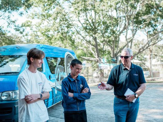 Three people stand talking outside near a blue van on a sunny day. Two young men are smiling, while an older man in sunglasses gestures with a pen and holds a notepad. Trees and fencing are visible in the background.
