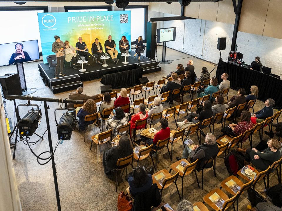 A diverse audience watches a “Pride in Place” panel discussion on stage, with six speakers seated and one standing. An Auslan interpreter is visible on a screen to the left. The room is brightly lit and modern.