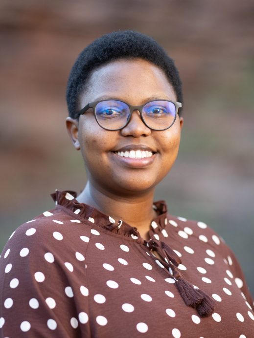 A person with short hair and glasses smiles at the camera. They are wearing a brown top with white polka dots and standing in front of a blurred outdoor background.