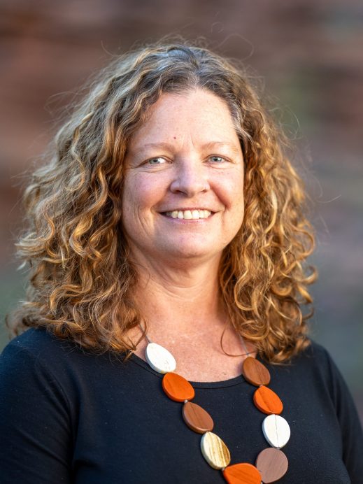 A woman with curly, shoulder-length light brown hair smiles at the camera. She is wearing a black top and a necklace with large round beads in orange, white, and beige. The background is softly blurred.