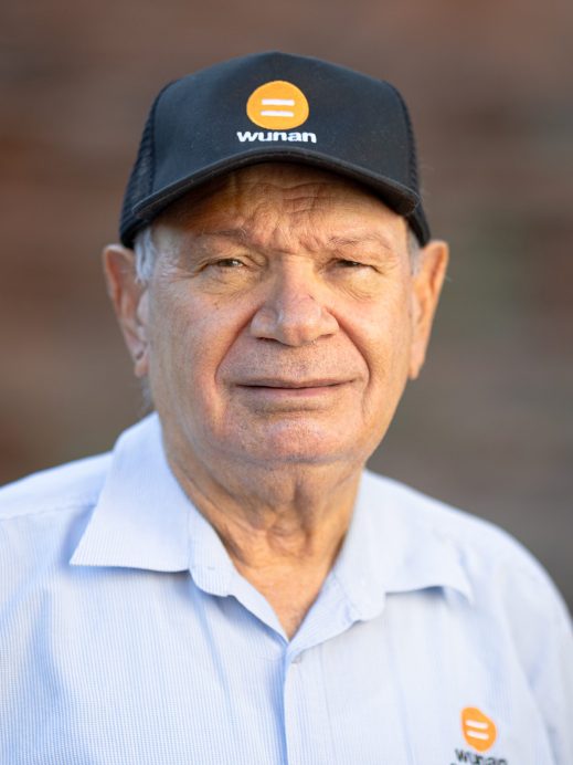 An older man wearing a black Wunan cap and a light blue Wunan shirt, standing outdoors and looking at the camera with a slight smile. The background is blurred.