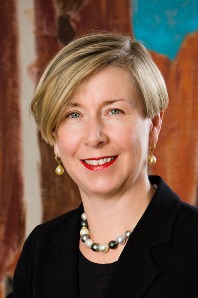 A woman with short blonde hair, wearing a black blazer, pearl necklace, pearl earrings, and red lipstick, smiling in front of a colourful abstract background.