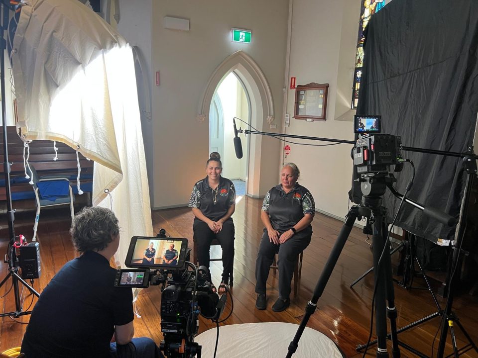 Two women sit side by side being filmed in a well-lit room with wooden floors. Cameras, lights, and microphones surround them, while a person films and interviews them. Stained glass windows are visible in the background.