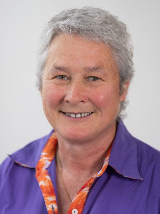 A smiling older woman with short grey hair wears a purple shirt with an orange-patterned collar, posed against a plain light background.