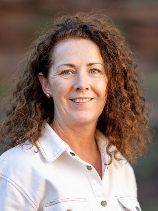 A woman with curly brown hair and blue eyes, wearing a light-coloured button-up shirt, smiles at the camera against a blurred outdoor background.