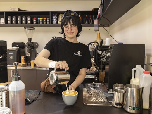 A barista wearing glasses and a black T-shirt pours steamed milk into a cup of coffee, creating latte art at a modern coffee shop counter with various barista tools and equipment about.