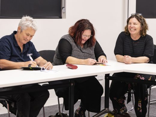Three women sit at a table in a meeting room. Two are writing and one is smiling. They appear to be engaged in a friendly discussion or collaborative activity.