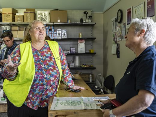 A woman in a yellow safety vest talks to another woman across a counter in a shop, with shelves of various items and two other people in the background.
