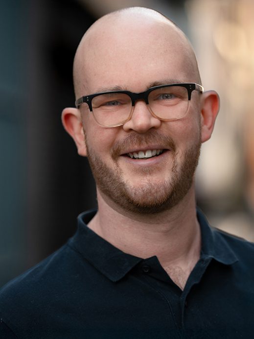 A smiling bald man with a beard and glasses wearing a dark collared shirt, standing outdoors with a blurred background.