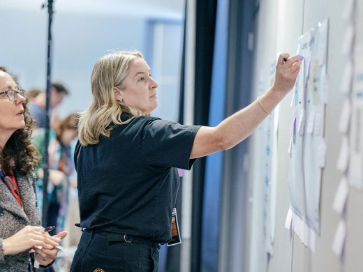 Two women stand by a wall covered with papers and sticky notes. One woman writes or adds a note to the wall, whilst the other observes. The background shows other people blurred out.