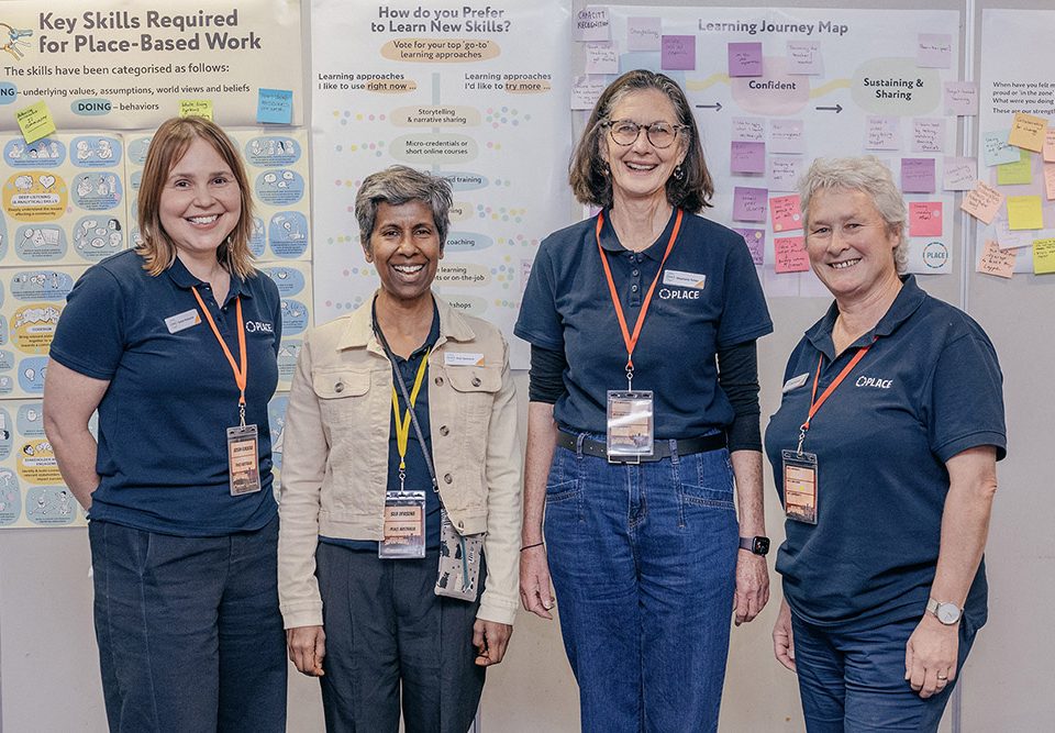 Four women stand smiling in front of posters about workplace skills and learning journeys. They wear name badges and navy blue shirts, suggesting they are part of the same team or organisation.