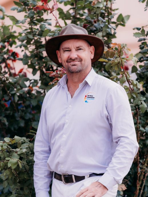 A man wearing a wide-brimmed hat, light purple button-up shirt, and white trousers stands in front of greenery and red flowers, smiling with one hand resting by his side.