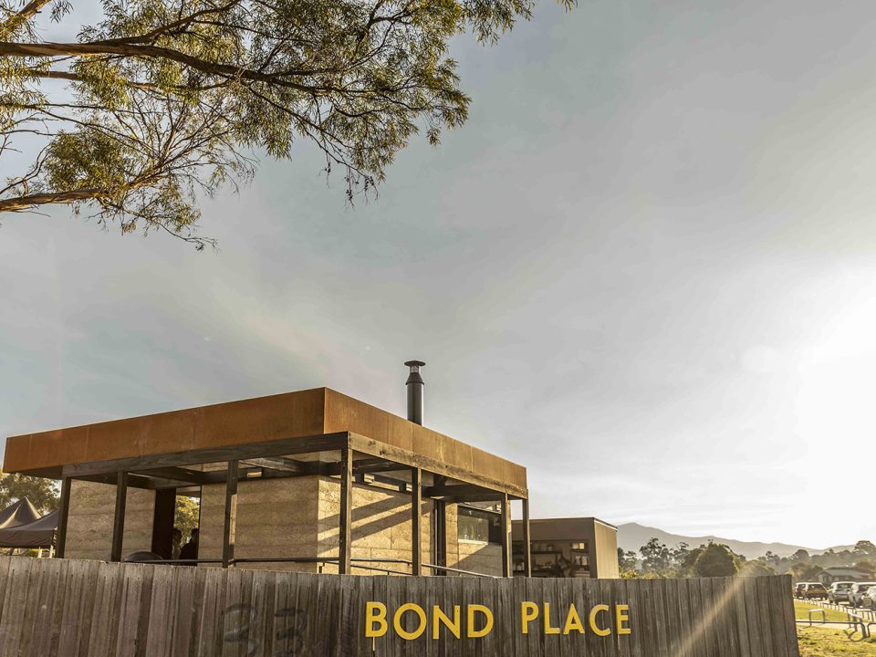 A modern building with a rust-coloured metal roof and chimney sits behind a wooden fence with yellow letters spelling BOND PLACE. Trees, cars, and mountains are visible in the background under a clear sky.