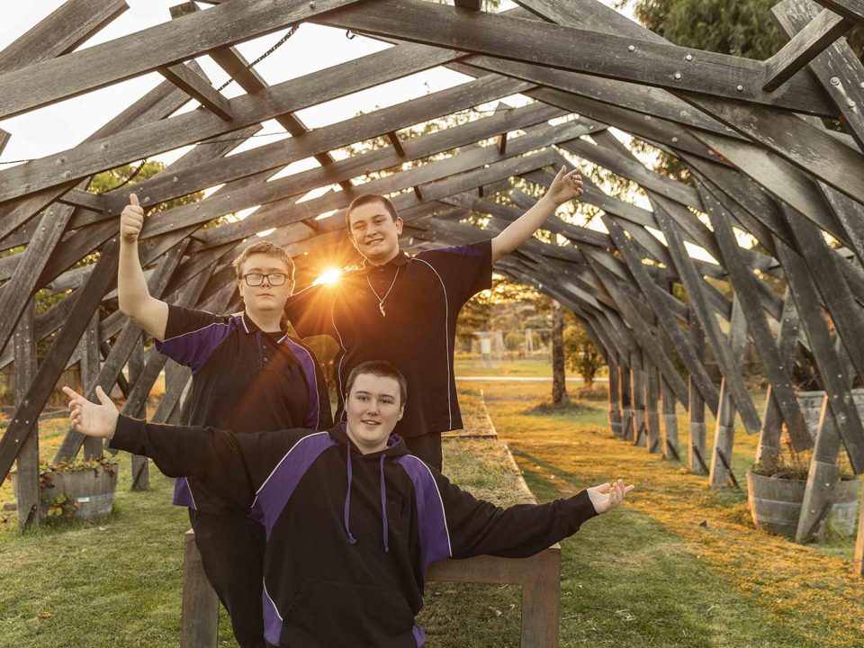 Three teenagers wearing black and purple hoodies stand under a wooden pergola at sunset, smiling with arms outstretched. Sunlight shines through the structure, illuminating the grassy area behind them.