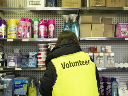 A person wearing a yellow Volunteer vest stands in front of shelves stocked with toiletries and household supplies in a shop or donation centre.