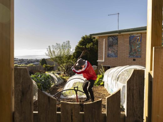 A person wearing a red jacket and hat is working with a garden tool in a back garden, surrounded by raised beds covered with plastic near a brick house on a sunny day.