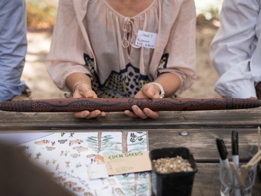 A person wearing a name badge holds a decorated wooden stick with carved patterns over a table with papers, a seed packet, seeds, and pens. Other people are partially visible beside them.