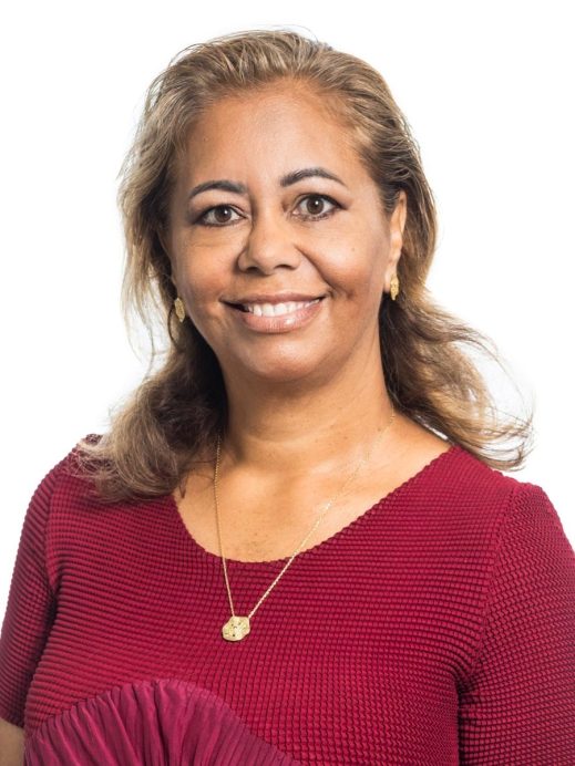 A woman with light brown hair and medium skin tone smiles at the camera. She is wearing a textured burgundy dress, gold earrings, and a gold necklace, posing in front of a plain white background.
