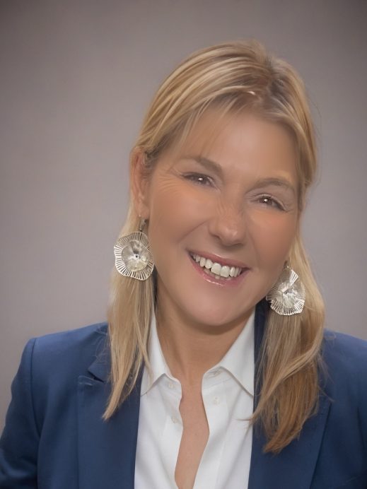 A smiling woman with blonde hair, wearing a blue blazer, white shirt, and large silver circular earrings, poses in front of a neutral grey background.