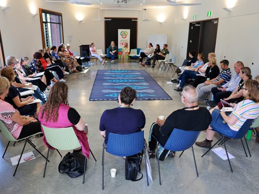 A group of people sit in a large circle on chairs in a bright room, engaged in a discussion or workshop. Some hold notebooks or coffee cups; a patterned rug lies in the centre of the circle.