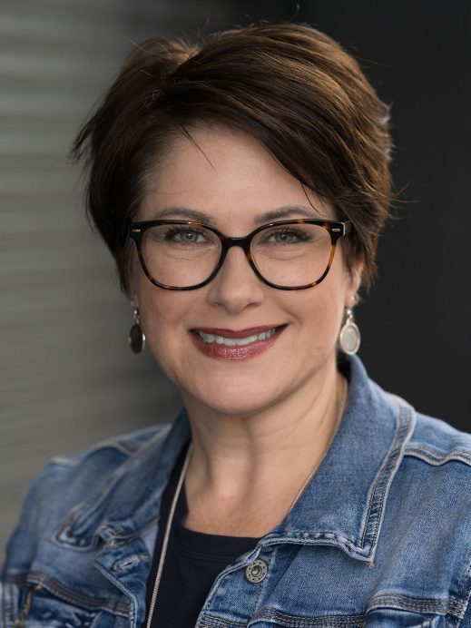 A woman with short brown hair and glasses smiles at the camera. She is wearing a denim jacket, dark top, and oval earrings, with a blurred background of grey horizontal lines.