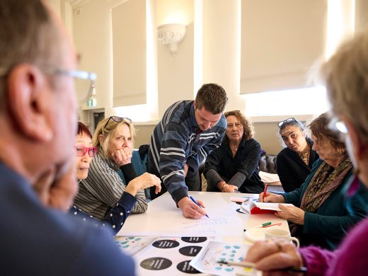A group of people are sitting around a table, all looking and listening to one person close to the camera. There are different pieces of paper on the table, and one person is writing notes on a large piece of paper.