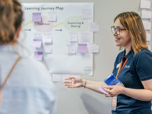 A woman in a navy PLACE polo shirt is explaining to a group of people in front of her while gesturing towards a poster on the wall titled 'Learning Journey Map'. It has lots of post-it notes stuck on it.