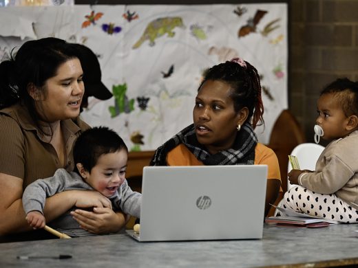 Two women are talking and sitting in front of a laptop. The woman on the left is wearing a brown top and holding a toddler who is playing with wooden sticks. The woman on the left is wearing a scarf and an orange top, and there is another toddler next to her sitting on the table.