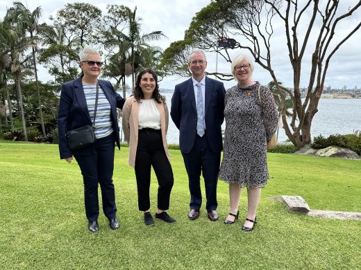 Four people are standing in a line outside on grass. Behind them are multiple trees and the water of Sydney Harbour.